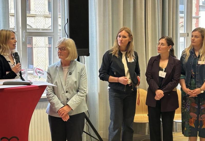 Fünf Frauen stehen in einem großen Raum an einem Rednerpult und hören einer Ansprache zu