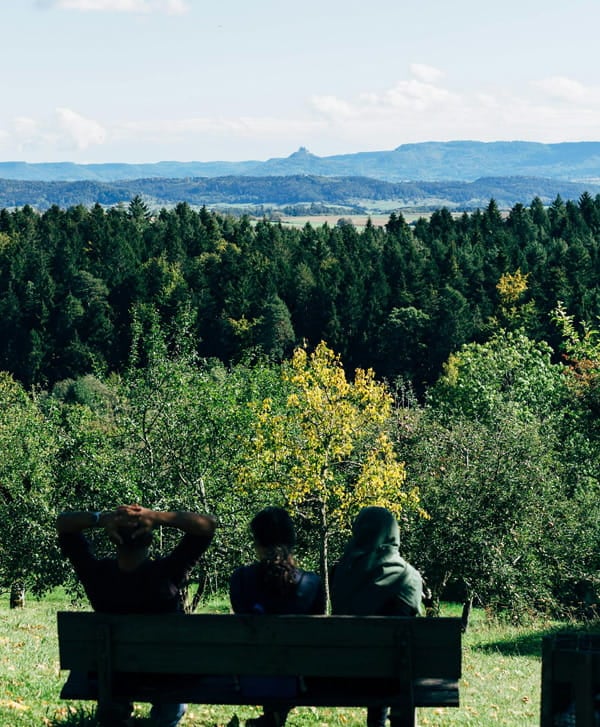 Gemeinschaft mit Ausblick ins Grüne Drei Menschen sitzen auf einer Bank und blicken über Wald und Landschaft in die Ferne