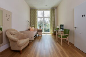 Helles Bewohnerzimmer im Robert Krekel Haus mit Pflegebett, Sessel, Tisch, Fernseher und großem Fenster mit Balkonzugang