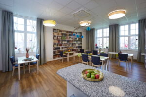 Gemeinschaftsraum im Robert Krekel Haus mit Tischen, Stühlen, Bücherwand und heller wohnlicher Atmosphäre