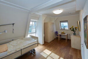 Freundlich eingerichtetes Bewohnerzimmer im Dachgeschoss des Robert Krekel Hauses mit Pflegebett, Kleiderschrank und Sitzplatz