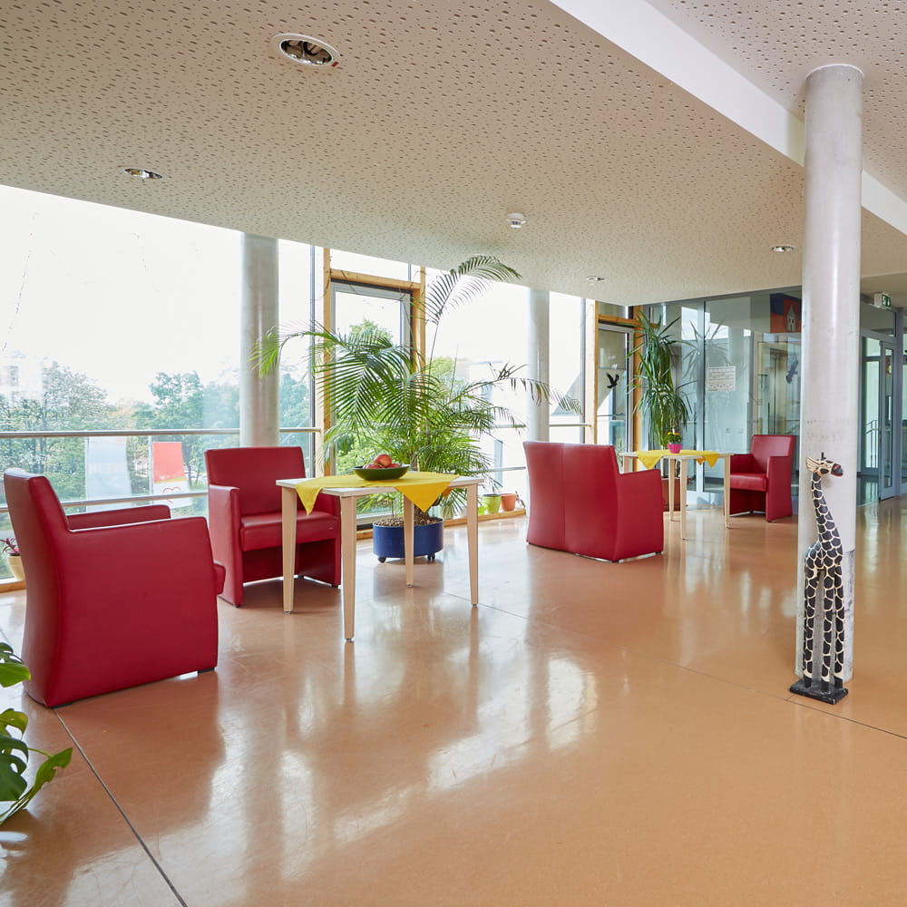 Großzügiger, lichtdurchfluteter Aufenthaltsbereich mit roten Sesseln, großen Fensterfronten und direktem Zugang zur Dachterrasse der neurologischen Fachpflege