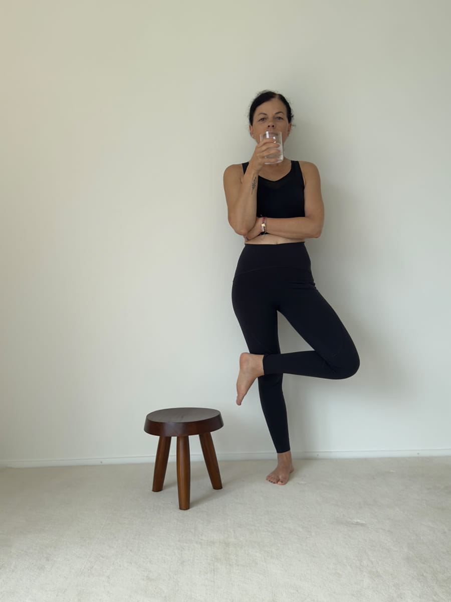 Yogalehrerin in schwarzer Sportkleidung steht auf einem Bein neben einem Holzhocker und hält ein Glas Wasser